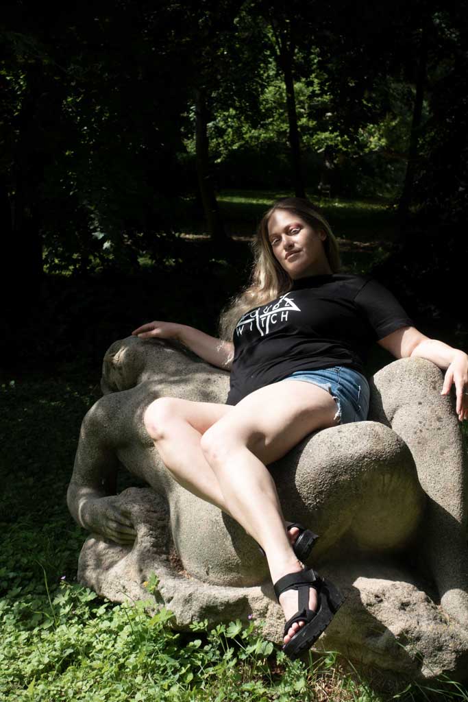 Artist Denise Kottlett sitting on a stone statue depicting a woman wearing With House, a black 4XL T-Shirt by House of the House.