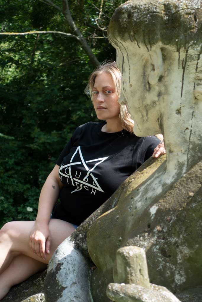 Artist Denise Kottlett sitting behind a stone statue of a female head wearing Witch House, a black 4XL T-Shirt by House of the House.